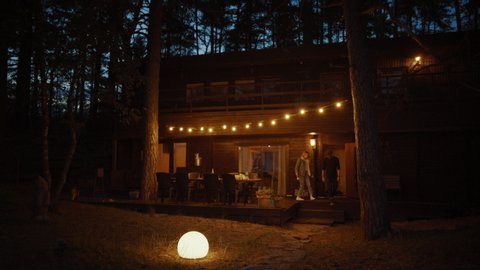 Young Man and Woman Walk From a Cozy Country House Together. House has Warm Lights Hanging Above the Terrace. Romantic Summer Evening Atmosphere. Cottage is Situated in a Pine Forest.