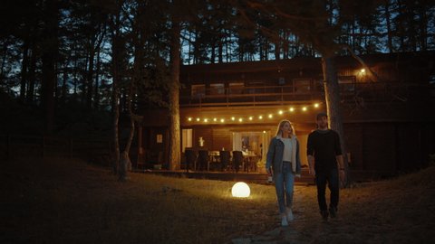 Happy Young Man and Woman Walk From a Cozy Country House Together. House has Warm Lights Hanging Above the Terrace. Romantic Summer Evening Atmosphere. Cottage is Situated in a Pine Forest.