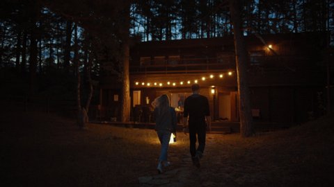 Young Man and Woman Walk to a Cozy Cottage Together. House has Warm Lights Hanging Above the Terrace. Romantic Summer Evening Atmosphere. House is Situated in a Pine Forest.