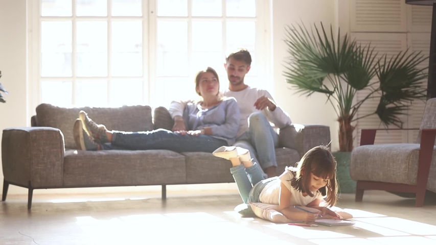 While little cutie daughter lying on warm floor with underfloor heating using colored pencils drawing on album young parents resting on couch, full family spending weekend at modern comfy home concept Royalty-Free Stock Footage #1034755961
