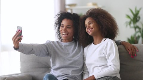 Different ages females young mixed race mother and grown up daughter adolescent smiling take self portrait selfie picture, relative people sit on couch at home using gadget having fun with modern tech