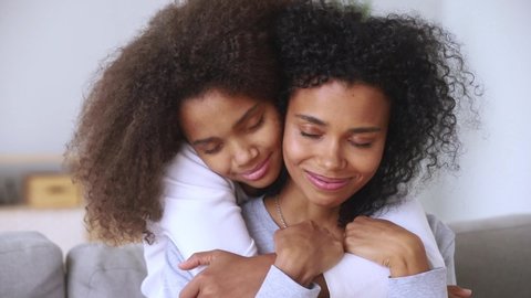 Close up view attractive afro biracial mother and grown-up pre-teen daughter or different ages sisters embracing on couch closed eyes enjoy moment of tenderness, love protection and gratitude concept