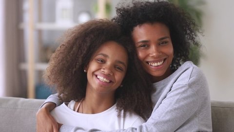 Mixed race mommy cuddle teenage daughter, different age sisters smile pose looking at camera sit on couch in living room head shot, relatives people, warm relations, understanding, mothers day concept
