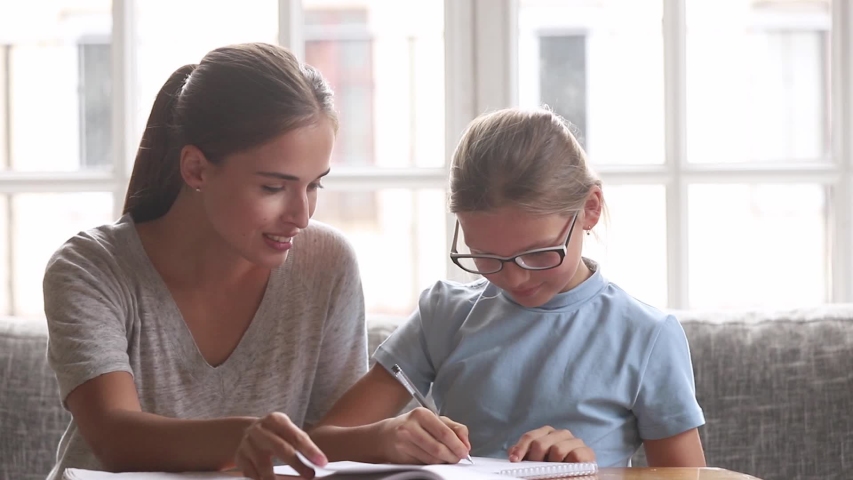 Mother helping to schoolgirl daughter with homework, babysitter, older sister or private tutor teaching schoolchild girl in glasses doing task together, educational process support learning concept Royalty-Free Stock Footage #1034756069