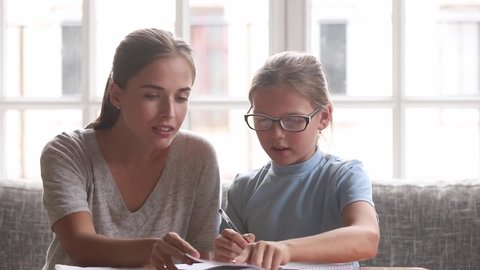 Mother helping to schoolgirl daughter with homework, babysitter, older sister or private tutor teaching schoolchild girl in glasses doing task together, educational process support learning concept