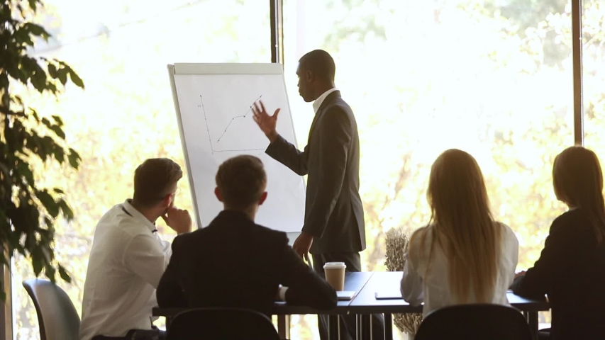 African coach stands near flip chart showing sales growth shown on raising graphic during corporate training finish presentation with positive forecast and participants applauses express gratitude Royalty-Free Stock Footage #1034756195