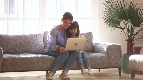 Mother daughter sitting on couch spending free time at home having fun together using laptop apps, browsing internet, watching or choose cartoons online, nanny teach kid electronic device use concept