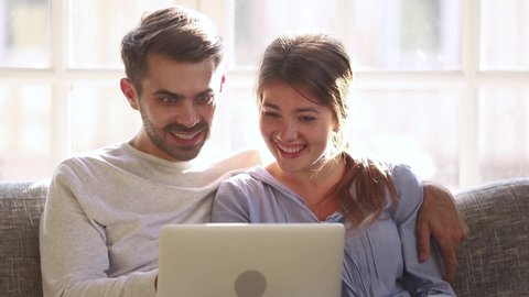 Couple sitting on couch browse internet offers read media news, husband embraces wife resting at home spend time use laptop feels surprised happy about cheap flight tickets travel opportunity concept
