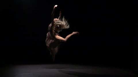 Slow motion of a sporty maiden in a beige dress dancing contemp in the darkness of a studio. She is performing the elements of dance against a black background in the beam of a spotlight.