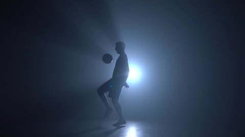 Silhouette of football freestyler stuffing the ball and making tricks in a twilight against a light of studio. Slow motion