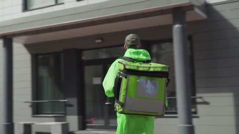 Tracking shot of young male food courier walking down street with insulated backpack using cell phone for navigation. Food delivery guy coming to house door, calling doorbell and speaking to customer