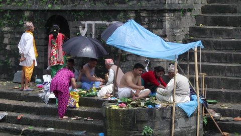 Pashupatinath , Kathmandu / Nepal - 04 27 2019: Group of people under umbrella and shanty shelter, eating & daily life at Pashupatinath riverside temple stairs. Poor poverty urban scene.