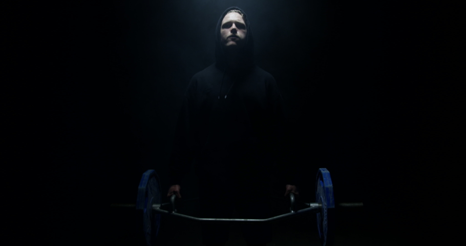 Male Weightlifter In Black Hoodie Looking At Camera And Lifting Weights In A Dark Background With Their Back To The Camera