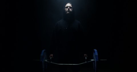 Male Weightlifter In Black Hoodie Looking At Camera And Lifting Weights In A Dark Background With Their Back To The Camera