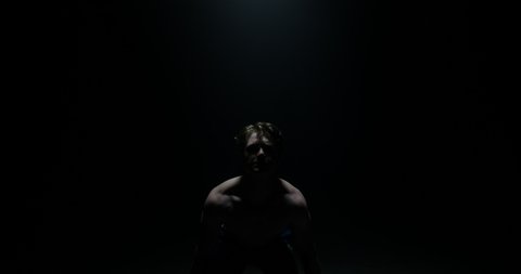 Shirtless Male Weightlifter In Black Hoodie Looking At Camera And Lifting Weights In A Dark Background With Their Back To The Camera