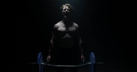 Shot Of Strong Shirtless Male Weightlifter Looking At The Camera And Lifting Weights In A Dark Background