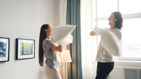 Cute couple is enjoying pillow fight then kissing and hugging on bed in light bedroom having fun in leisure time. Happy family, lifestyle and relationship concept.