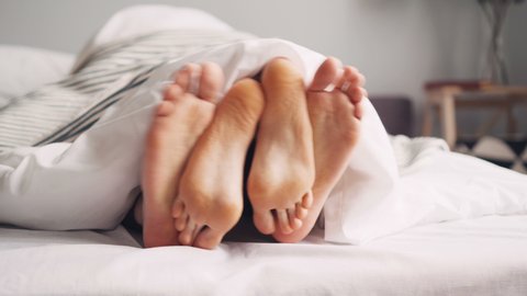Human feet are having fun in bed under blanket, girl is tickling guy moving legs on white sheets. Intimate relationship, family life and happiness concept.