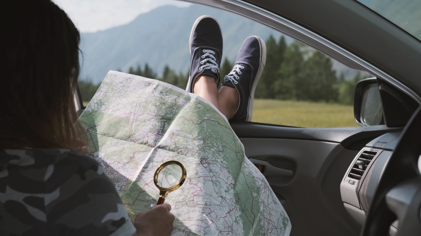 Girl tourist looks at a map using a magnifier while sitting in a car and sticking his legs out the window against the backdrop of the mountains