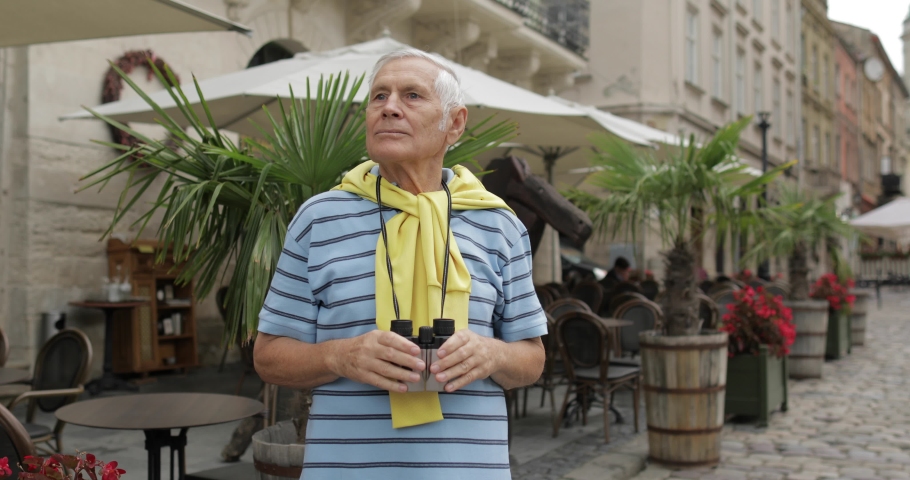 Senior male tourist exploring town and looking in binoculars. Traveling in Lviv, Ukraine. T-shirt and sweater in his neck. Vacation concept