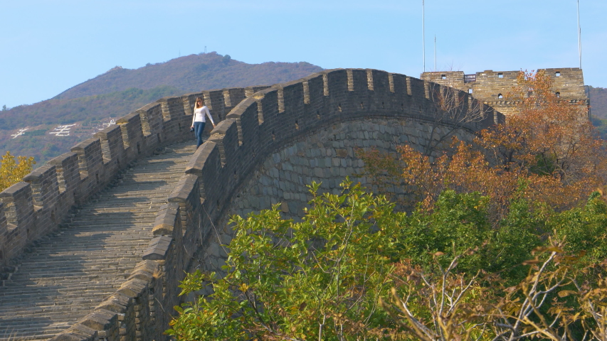 Young female photographer walks down the ancient stairs on top of the Great Wall of China on a sunny fall day. Woman walks along the ancient fortified stone wall crossing the green forests of China.