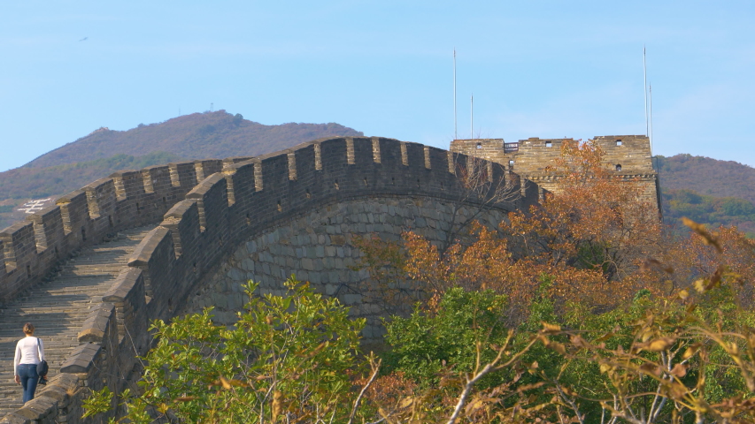 Unrecognizable female traveler walks up a flight of stairs atop the Great Wall of China on a sunny day. Woman walks along the walkway on top of the ancient stone wall crossing the forested mountains.