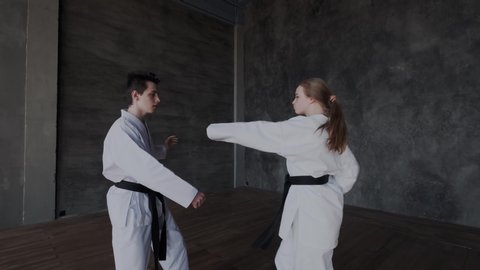 Slow motion of a dark-haired guy and a blonde girl while they are practice combat arts in komono with black belt. Sparring or duels in taekwondo class. Jump kick attack. Fight stance.