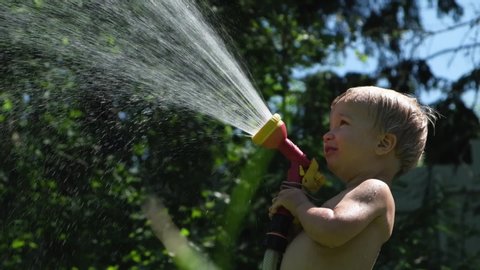 Cute toddler blond boy playing with garden sprinkler on summer hot day. Kid having fun on backyard spraying himself with a hosepipe. Happy childhood.
