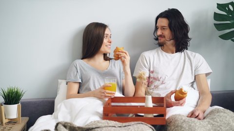 Joyful couple is eaing croissants in bed chatting and laughing enjoying morning in bedroom together. Modern lifestyle, relationship and people concept.