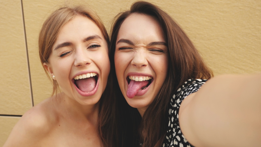Two young smiling hipster women in summer clothes.Girls taking selfie self portrait photos on smartphone.Models posing in the street near yellow wall.Female shows tongue, peace sign and make duck face