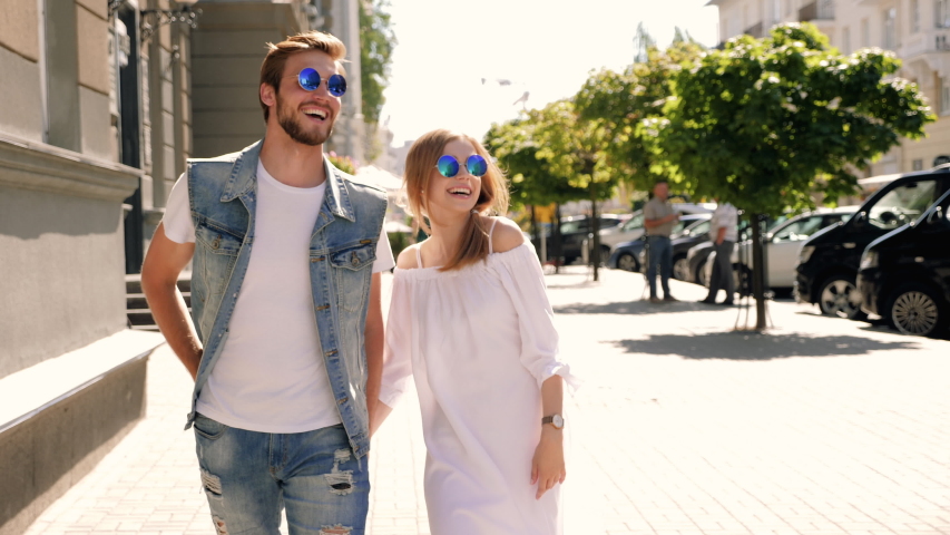 Smiling beautiful girl and her handsome boyfriend walking in the street. Woman in casual summer dress and man in jeans clothes.Cheerful couple family in sunglasses.Pointing on something interesting