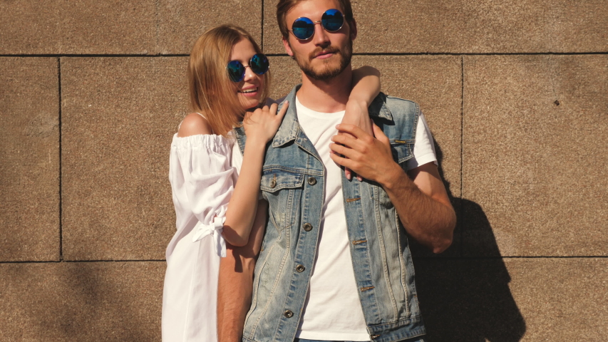 Smiling beautiful girl and her handsome boyfriend posing near wall in the street. Woman in casual summer dress and man in jeans clothes. Happy cheerful couple family having fun in sunglasses