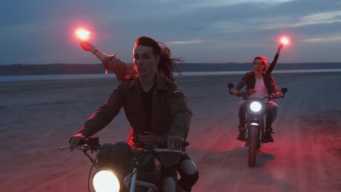 Two young couples riding on vintage motorcycles with red burning signal flares after sunset on beach, slow motion