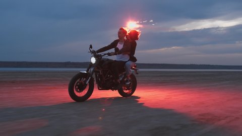 Young couple riding on vintage motorcycle with red burning signal fire after sunset on beach, slow motion