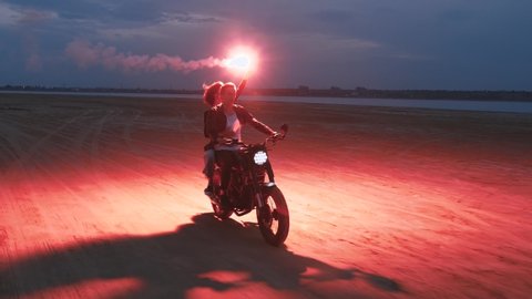 Couple riding on vintage motorcycle with red burning signal fire after sunset on beach, slow motion