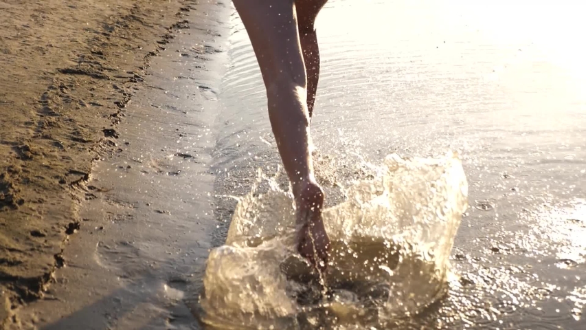 Bright sun. splashes of water fly in different directions. Slow motion. girl runs barefoot along shore and playing in the water on the beach at sunset. teen girl having fun in summer on the beach. | Shutterstock HD Video #1034865383