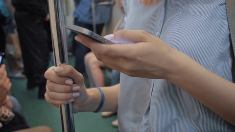 Close up of woman hand using smartphone apps or write messages and holding onto the railing in railcar with many passengers of underground subway, unrecognizable person.