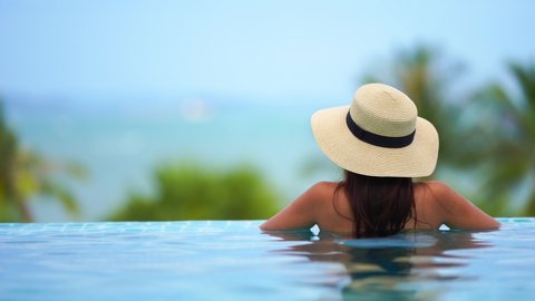 A woman with her back to the camera wearing a floppy Panama hat leans on the edge of a resort infinity pool looking out at the ocean horizon and palm trees.