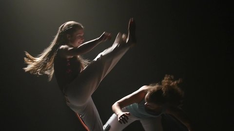 Close-up slow motion. Females are performing incredibly complex and steep movements of the martial art of capoeira in the twilight on a black background of studio. Acrobatic parkour.