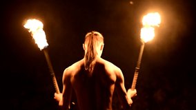 Close-up back view. Low key. Young male with long hair and bare torso rotates burning torch outdoors on a black night video slow motion. Modern fakir does tricks with a burning staff