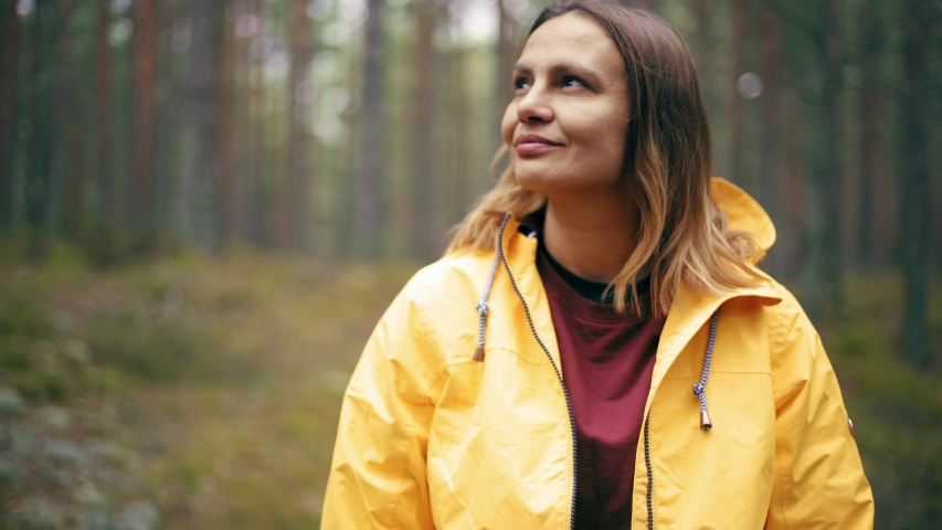 Portrait of a young woman in a bright yellow jacket standing at the rainy forest and looking around.