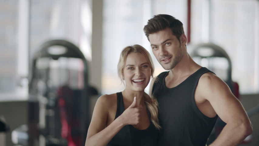 Happy fitness couple posing in modern gym. Smiling woman showing thumbs up gesture in fitness club. Satisfied woman showing like gesture after personal training in fitness center. | Shutterstock HD Video #1034920100