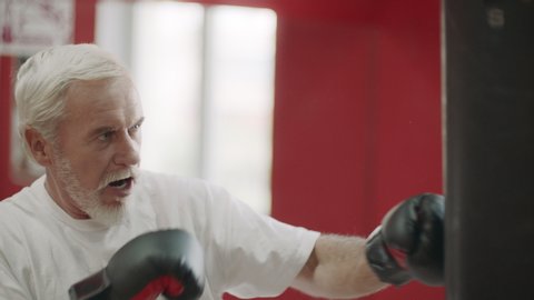 Old man training kick on boxing bag in fight club. Elderly man boxing punching bag at cardio training in fitness club. Portrait of handsome aged man boxing at fintess gym.