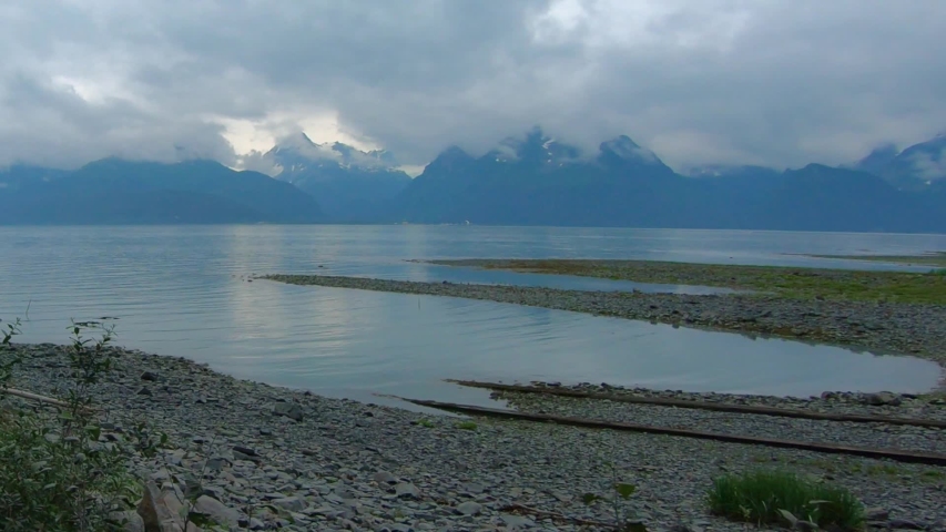 Passenger POV - driving by rocky beach of Resurrection Bay on Lowell Point with vehicles along the road and across the bay Kenai Mountains covered in a boreal forest; near Seward Alaska