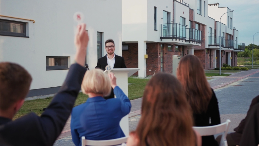 Fun auction sale. Back view of rich bidders raising hands bidding for real estate objetcs sitting outdoors. Charismatic male auctioneer leading a property auction.