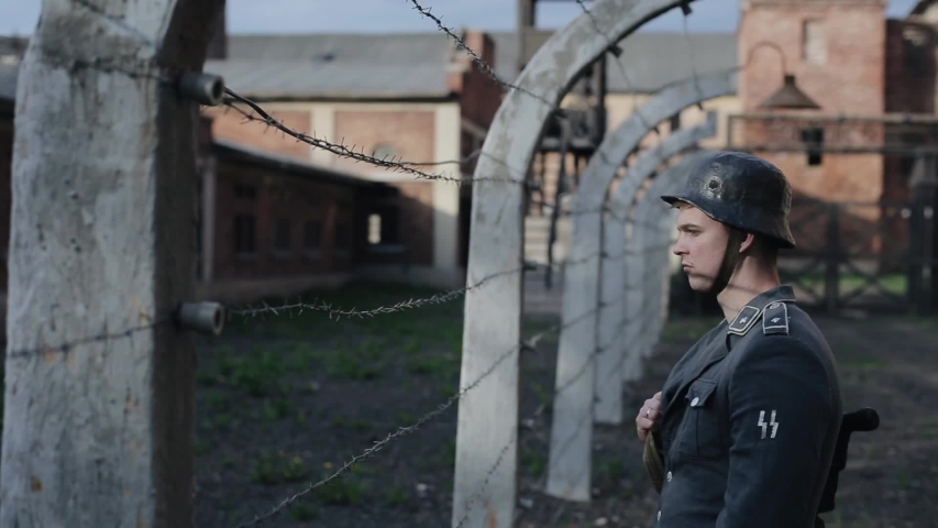 A close view of a young male actor dressed as a German soldier near a barbed wire fence guarding a death camp