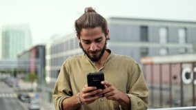 Smiling man texting by smartphone. Handsome bearded young man standing on street and using cell phone. Technology concept