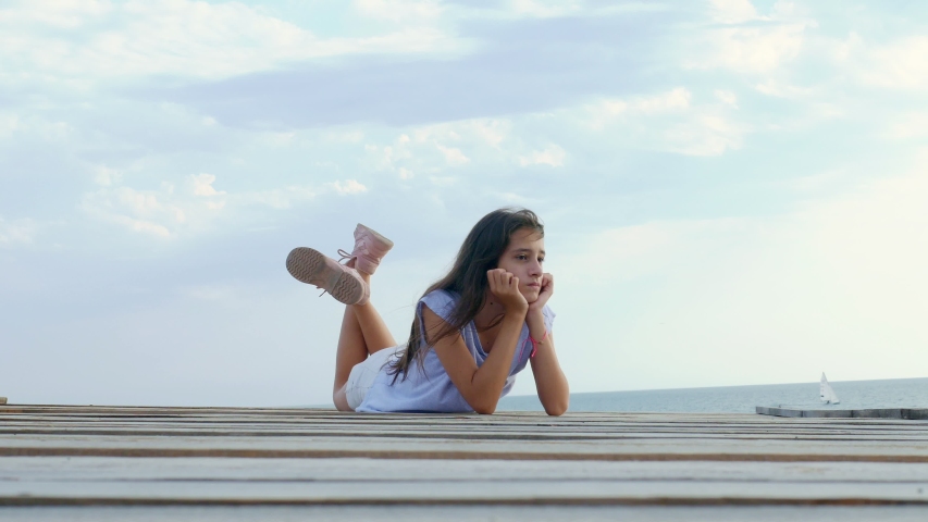 Teen girl sits alone on a wooden bridge at the sea and looks sad at the sea
