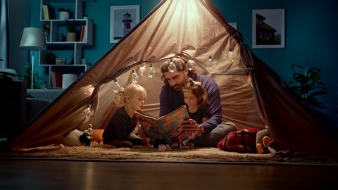 Happy dad and little daughters read a book in a tent at home