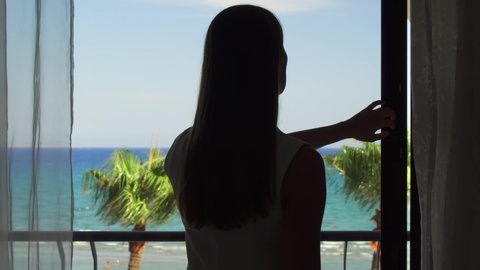 Young woman going out onto balcony and enjoying view from terrace. Female on vacation opening sliding balcony glass door and looking at Mediterranean sea. Wind swaying leaves on palm trees. Camera
